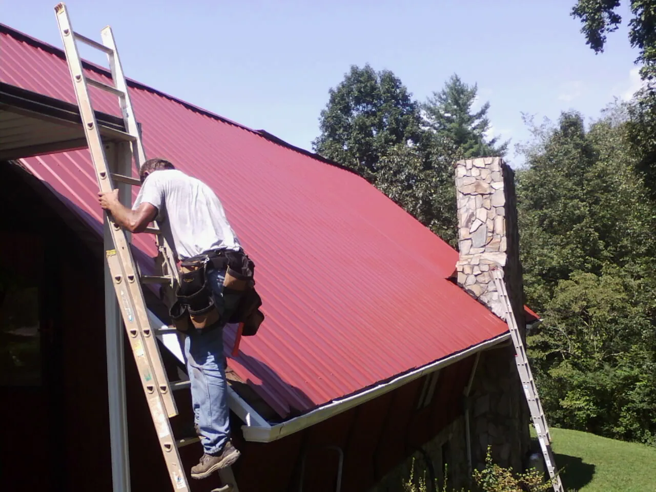 Roofer on a ladder beside a red metal roof and stone chimney — Soffit & Fascia Repair by Roofing Brothers in Uniontown